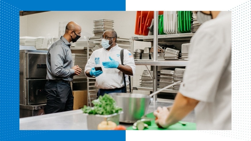 Set in a restaurant kitchen, a manager is seen speaking with an Ecolab associate who is wearing a mask and gloves while gesturing to something on his mobile or other small device. A kitchen worker cutting produce is blurred in the foreground.